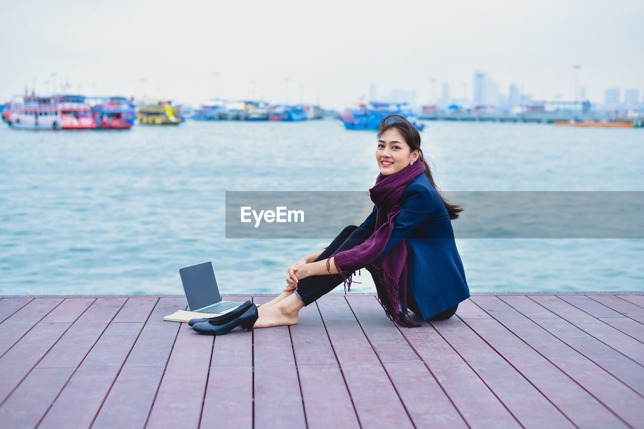 Full length portrait of smiling woman sitting on pier over sea