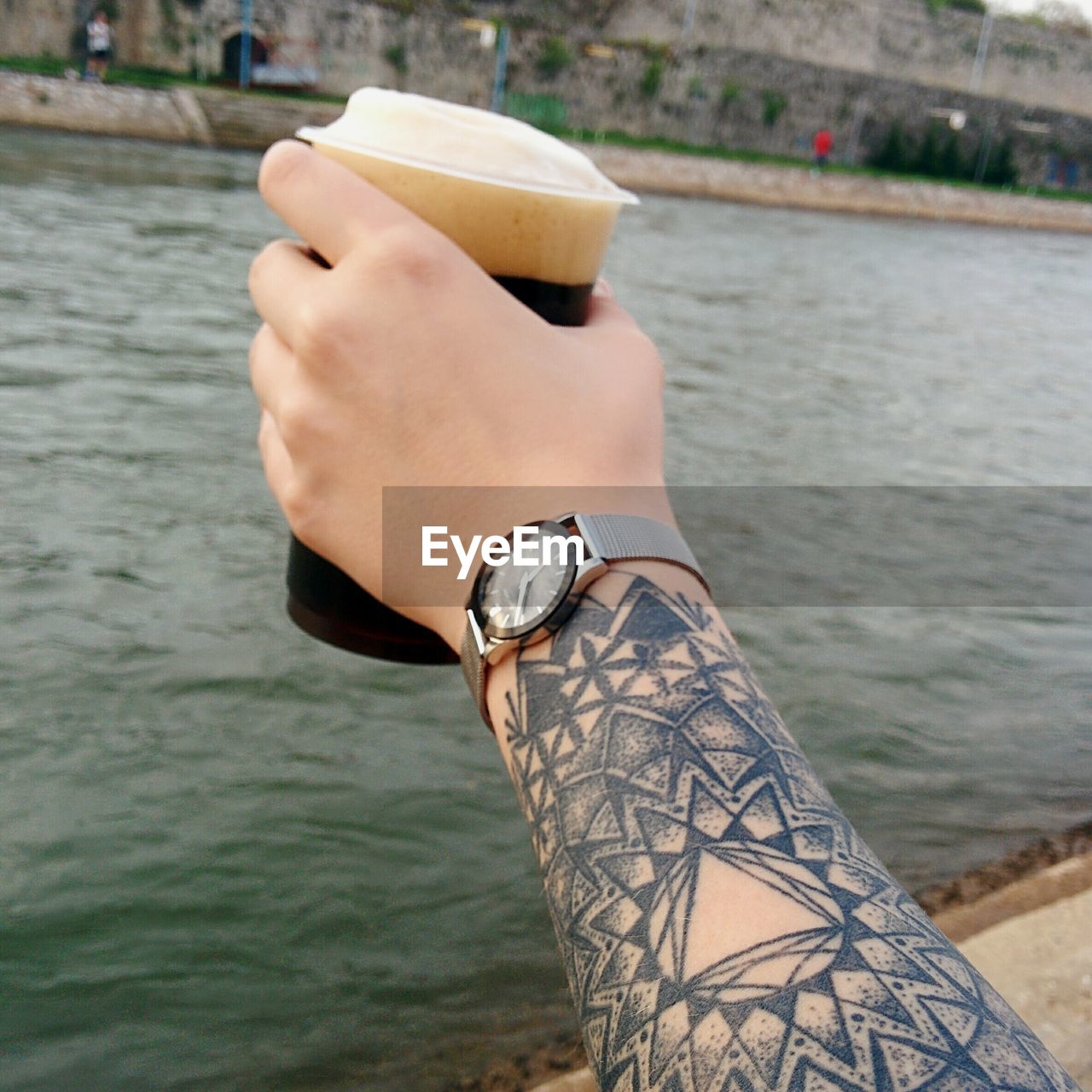 Cropped hand with mandala tattoo holding beer glass over river