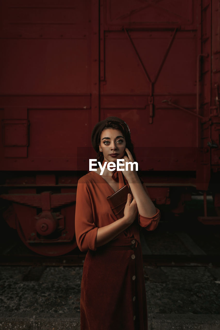 Dark haired female totally wearing terracotta clothes in vintage style standing near terracotta car train and holding open book in hands having dreaming look