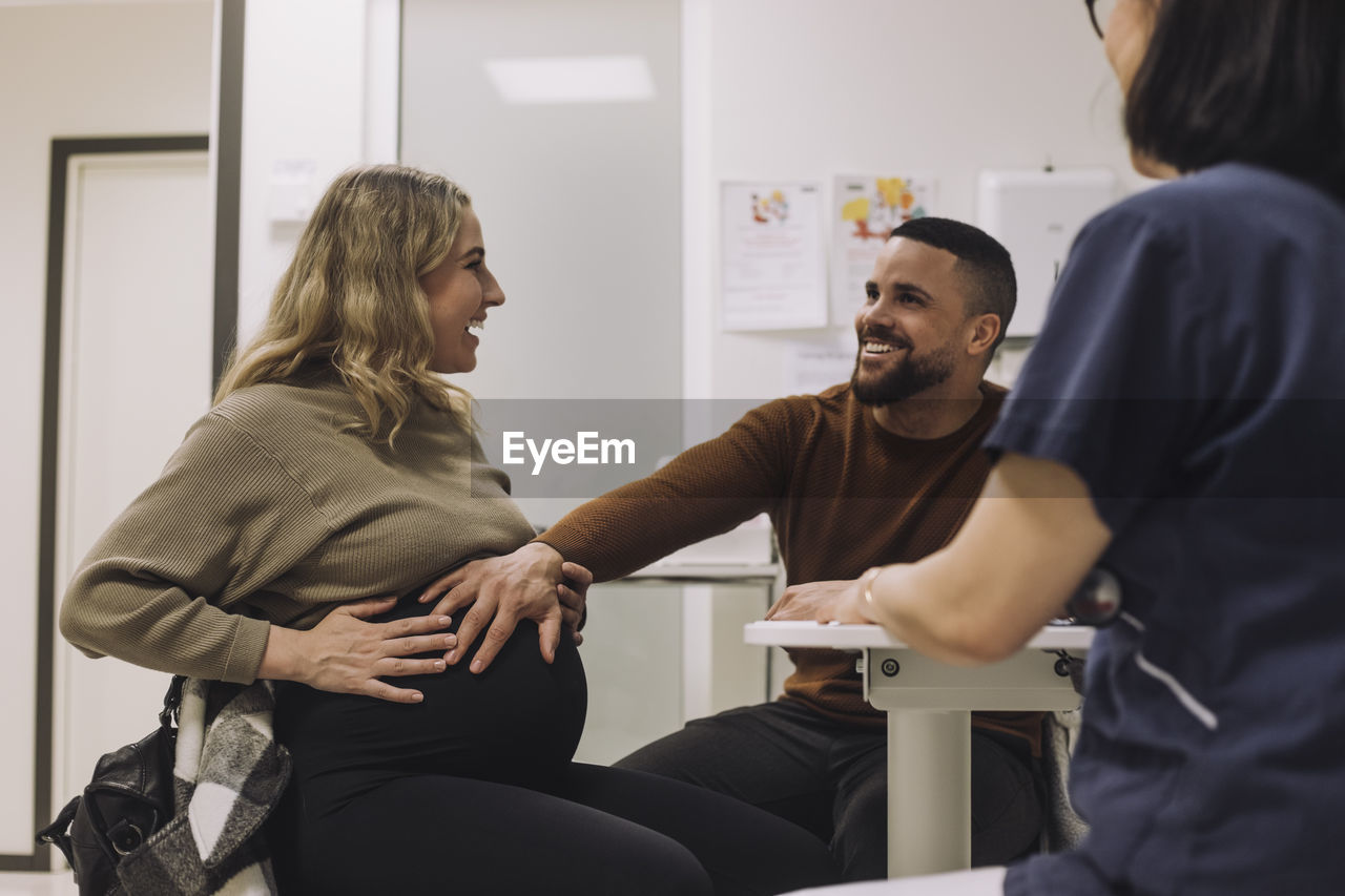 Happy man touching stomach of pregnant woman while sitting with female doctor in medical clinic