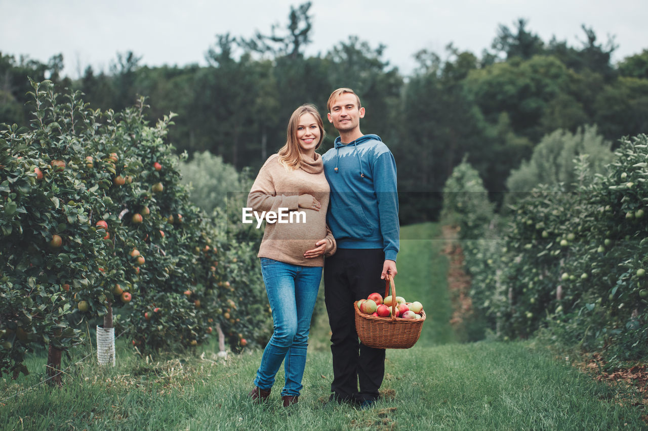 Portrait of smiling pregnant couple standing with apples in basket on grass