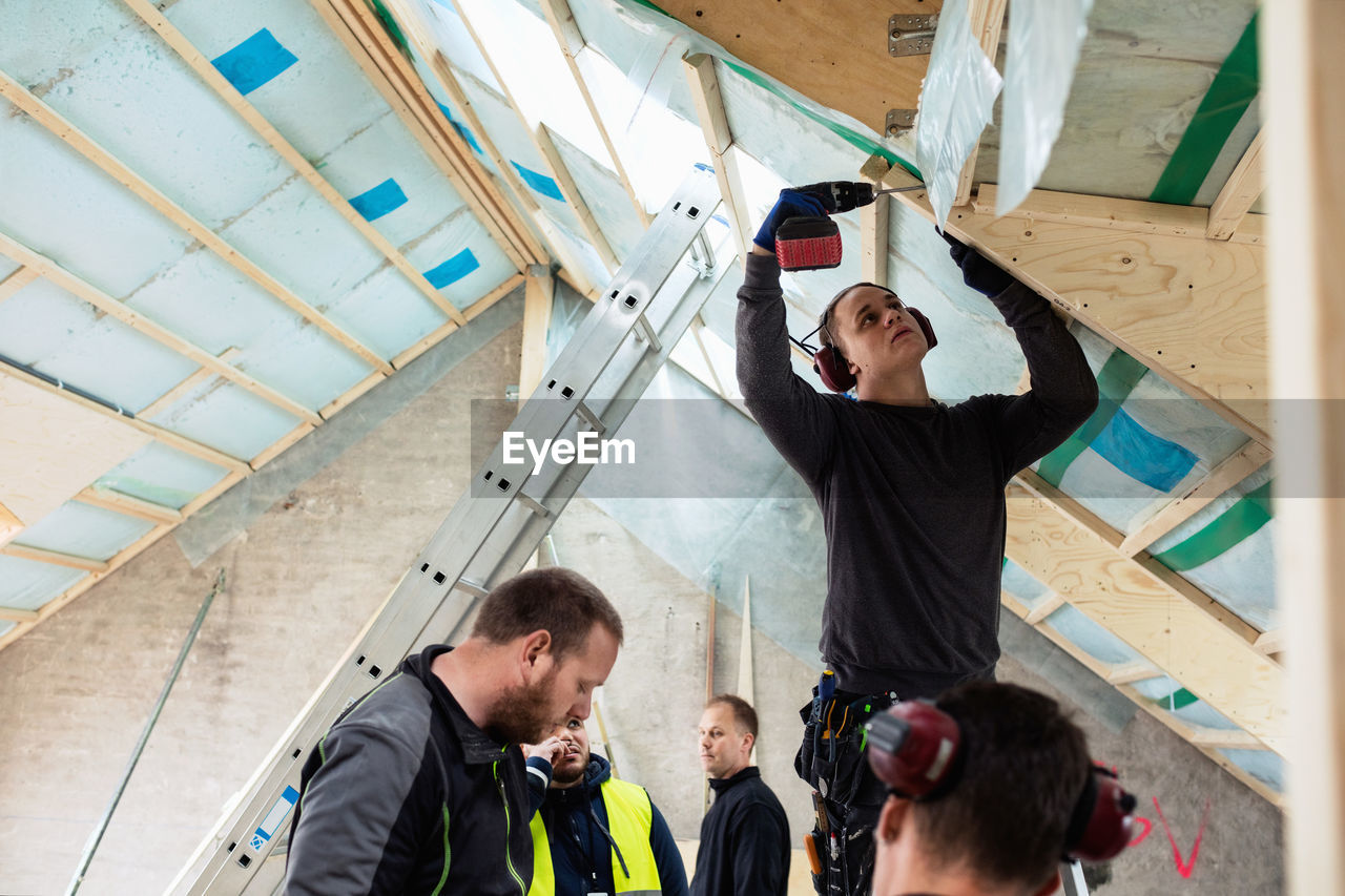 Construction worker drilling roof beam while coworkers standing by ladder at site