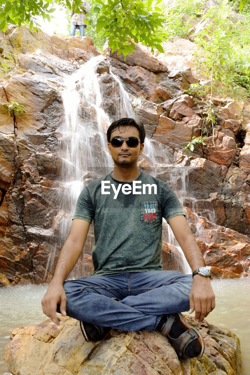 Portrait of young man sitting on rock against waterfall