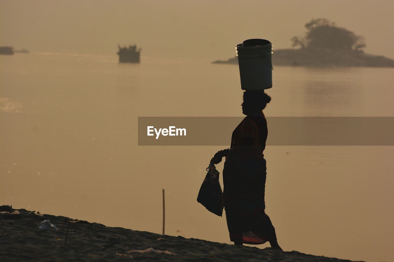 Silhouette woman with bucket on head walking at beach during sunset