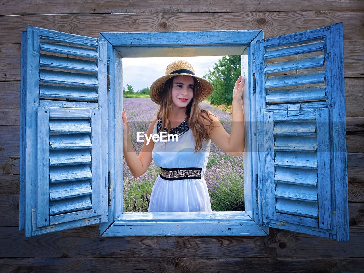 Woman wearing white dress and sun hat looking out through a wooden window