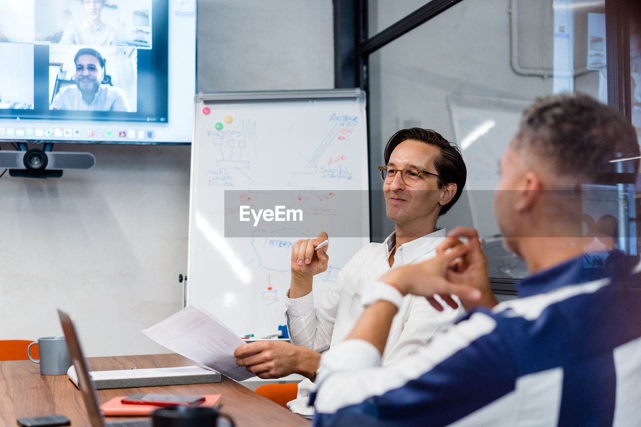 Coworkers sitting at table with gadgets and documents in modern workspace with white flip chat during online conference in office