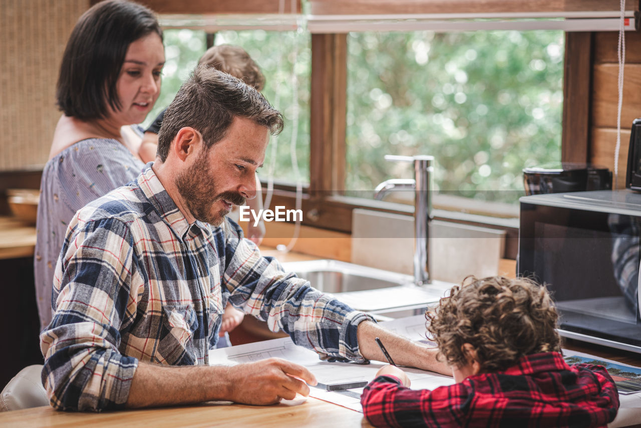 Side view of male architect sitting at table and creating blueprint of house in kitchen with family