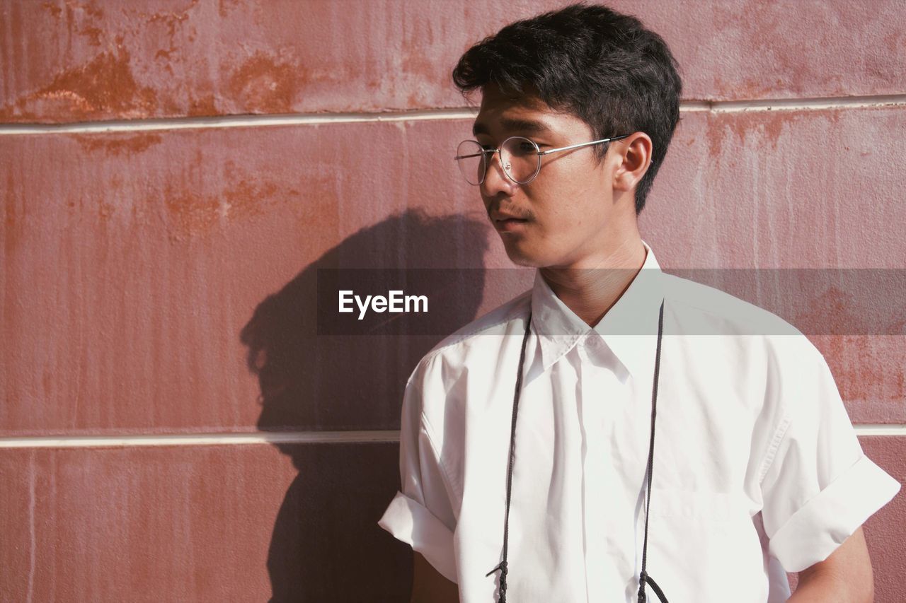 Thoughtful young man wearing eyeglasses standing against wall