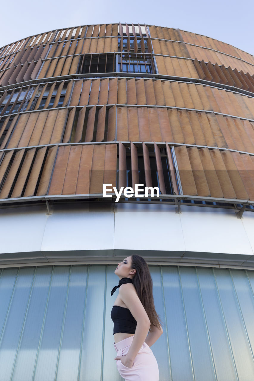 Fashionably dressed young woman looking at camera while standing near modern building in the city