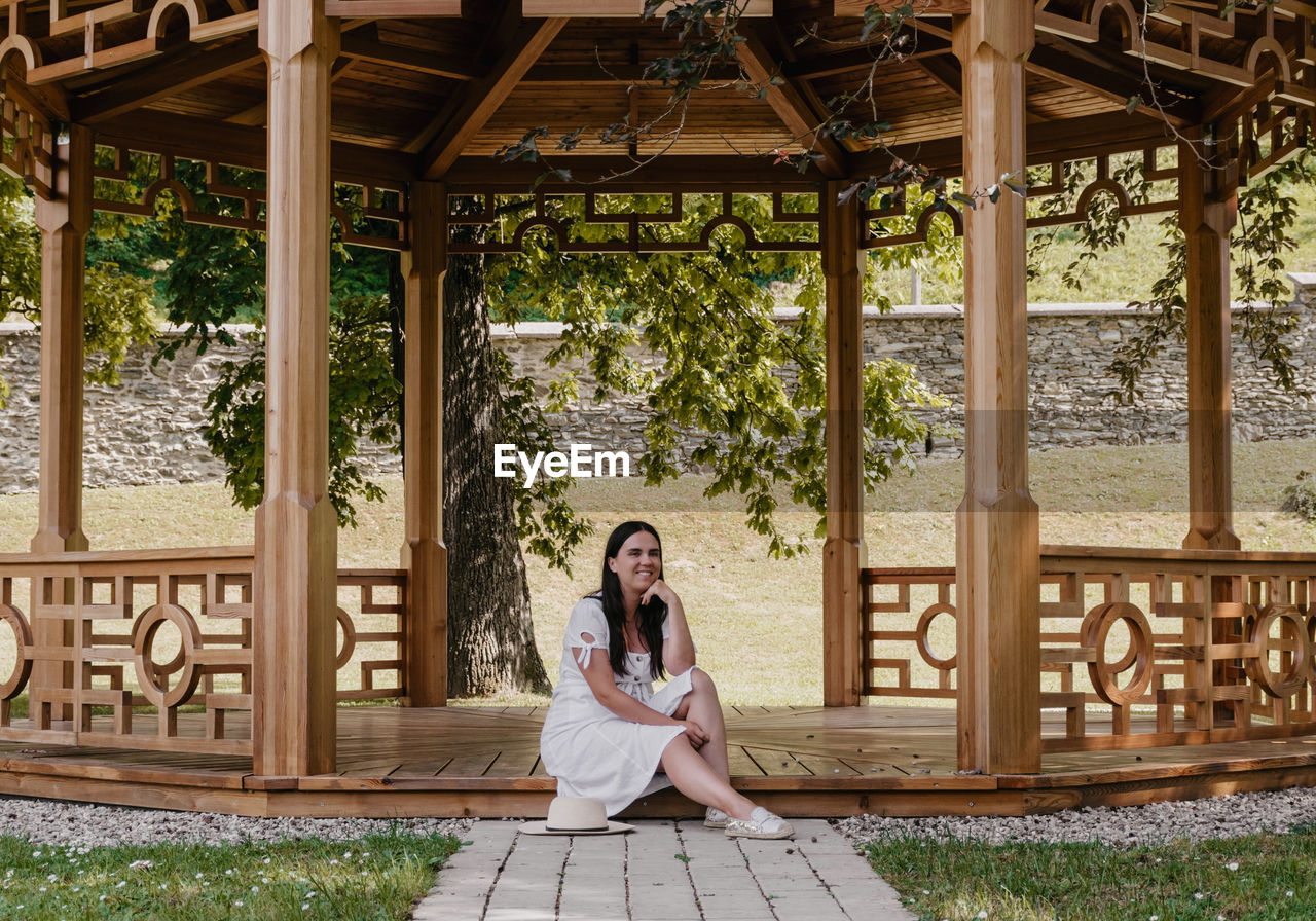 Portrait of beautiful young woman wearing white dress sitting under wooden pavilion in park.
