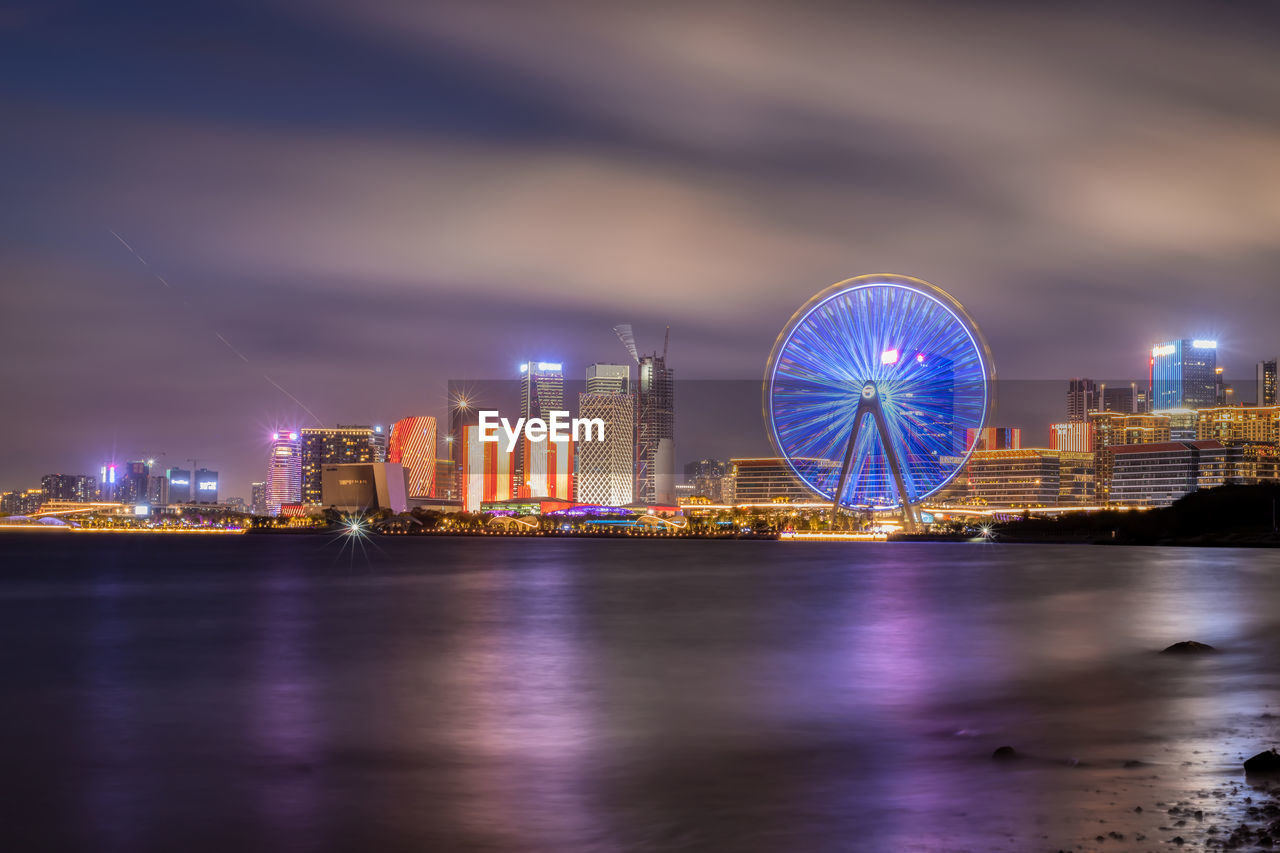 Illuminated ferris wheel by river and buildings against sky at night