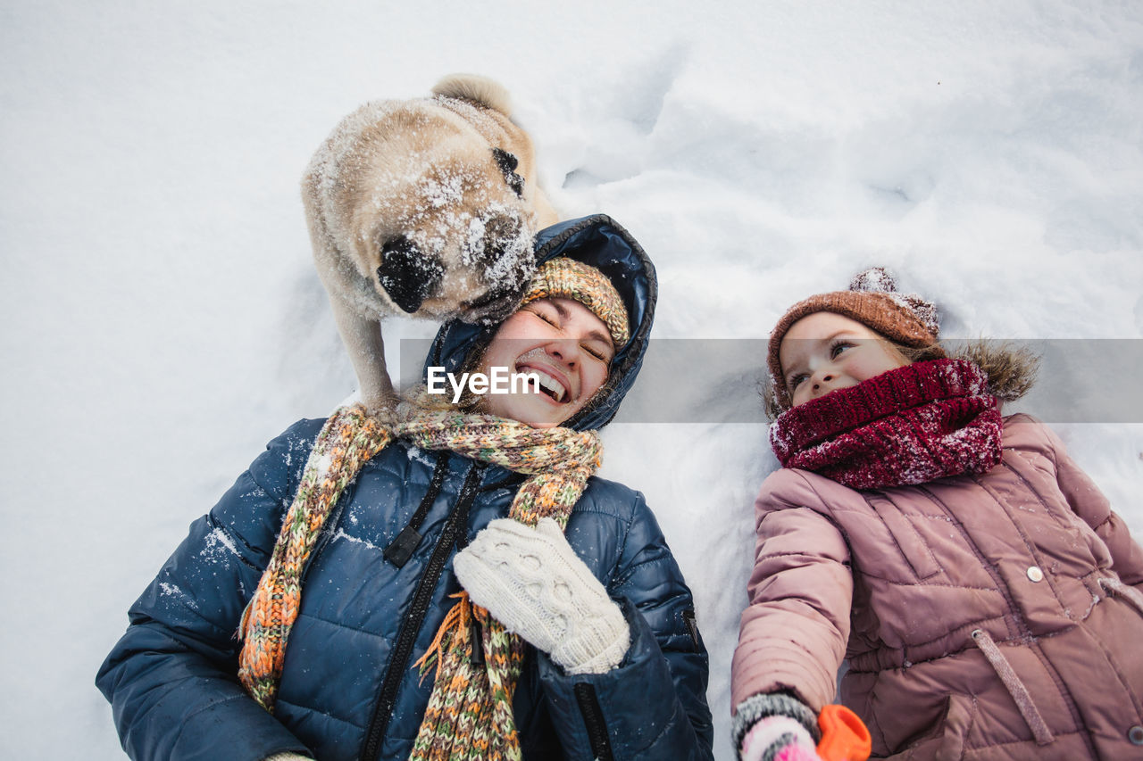 High angle view of dog licking cheerful mother lying with daughter on snow