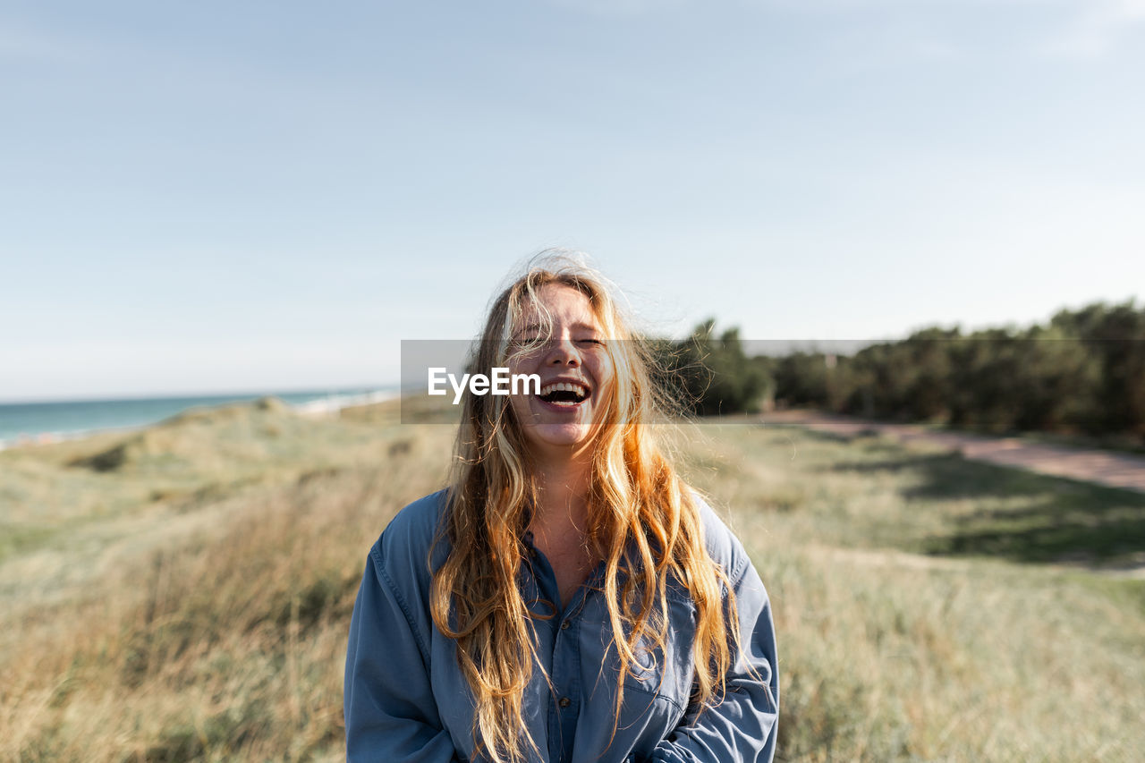 Cheerful young female in casual outfit standing on grassy meadow in summer with eyes closed