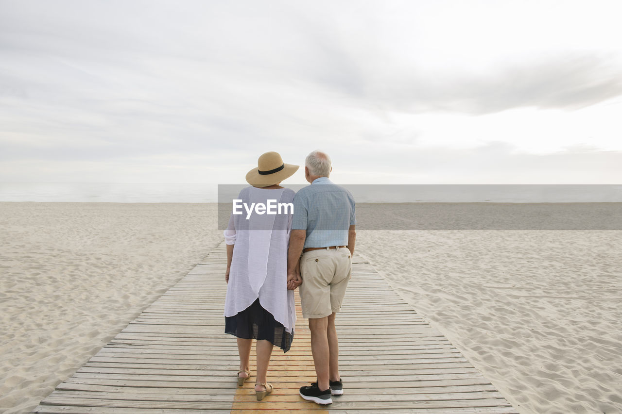 Back view of senior couple standing hand in hand on wooden boardwalk looking at horizon, liepaja, latvia
