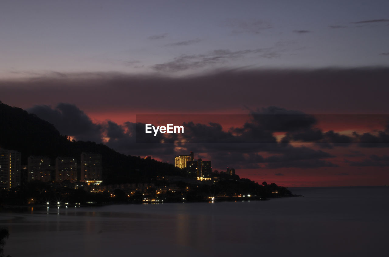 Illuminated city by sea against sky at sunset