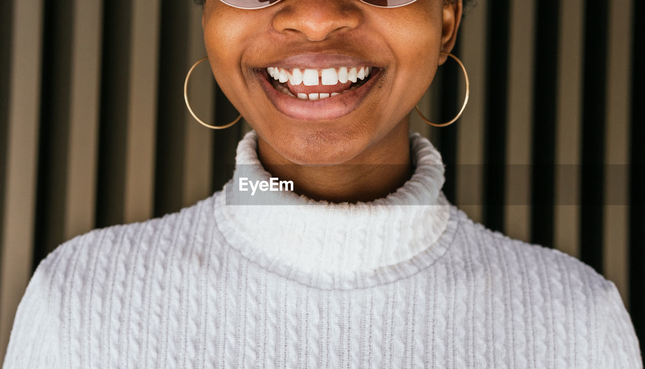 Crop african american female with gapped teeth in trendy sweater smiling happily against striped wall on street