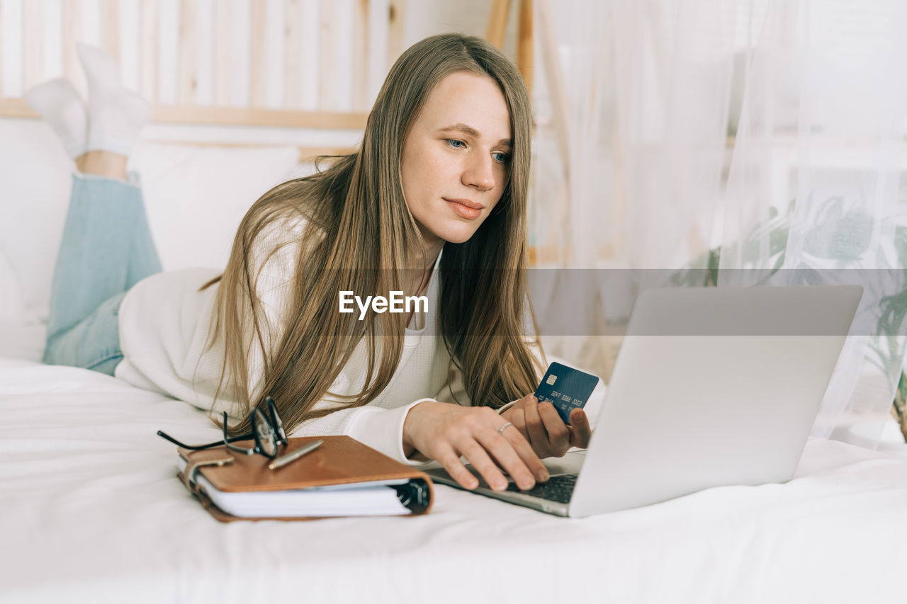 Freelance woman with credit card and notebook typing at laptop and online shopping, lying on the bed