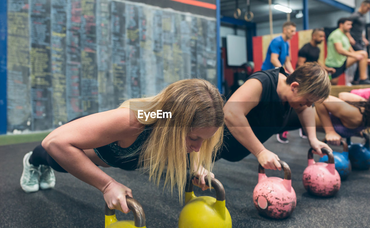 Sportswomen doing push-ups with kettlebells with mates doing box jumps in background