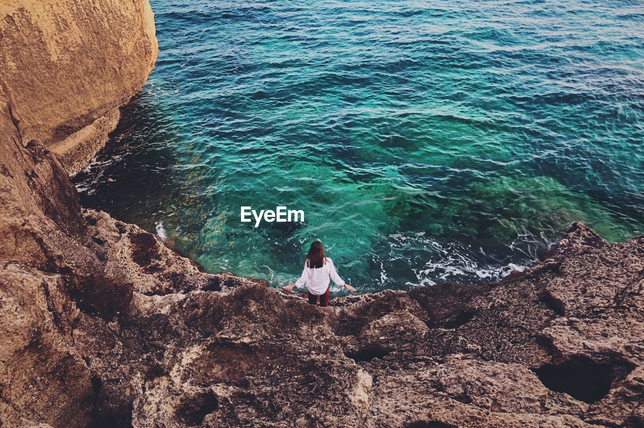 HIGH ANGLE VIEW OF WOMAN SITTING ON ROCK AT SEA