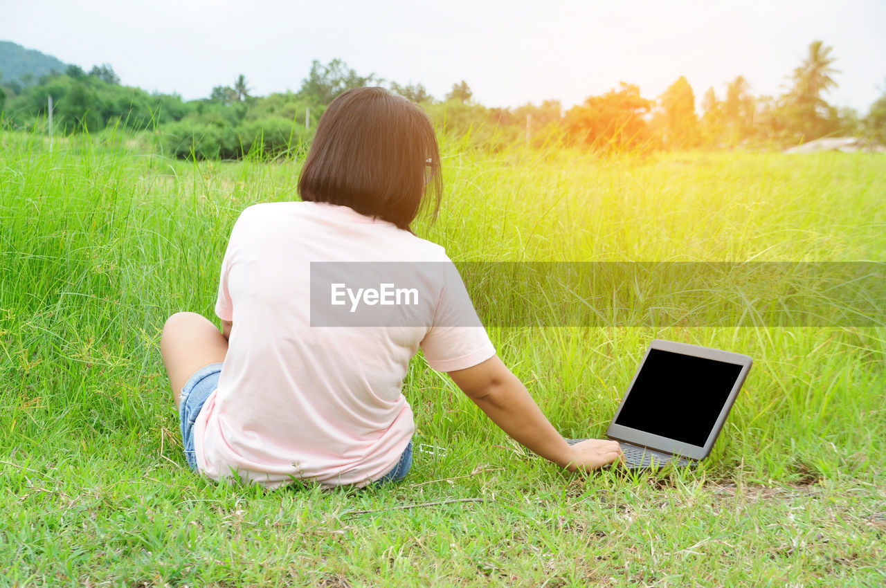 Rear view of woman using laptop while sitting on grassy field