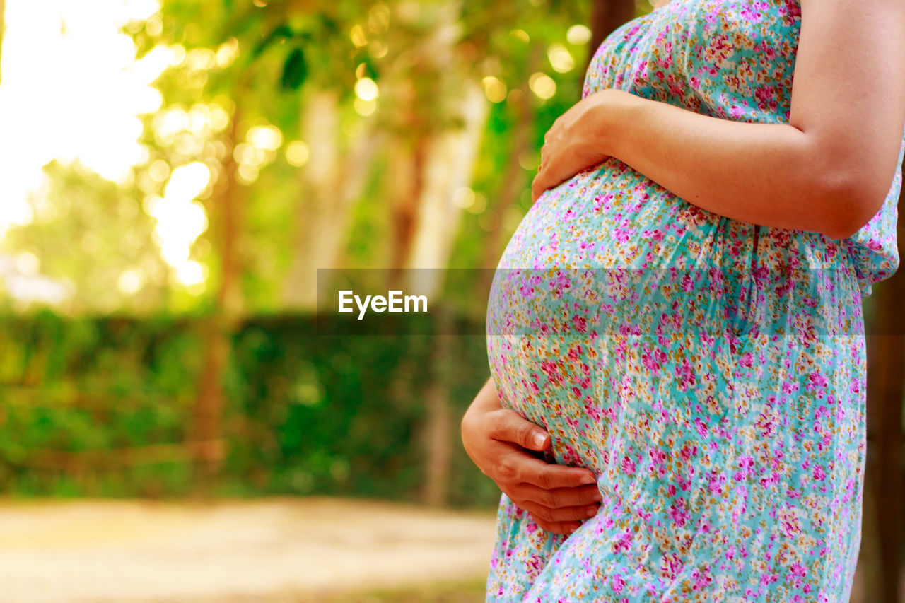 Midsection of pregnant woman touching abdomen while standing against trees