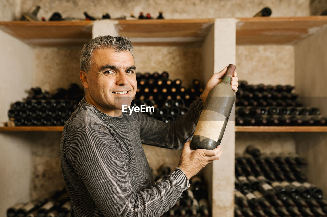 Smiling male owner holding old wine bottle while standing by rack at cellar