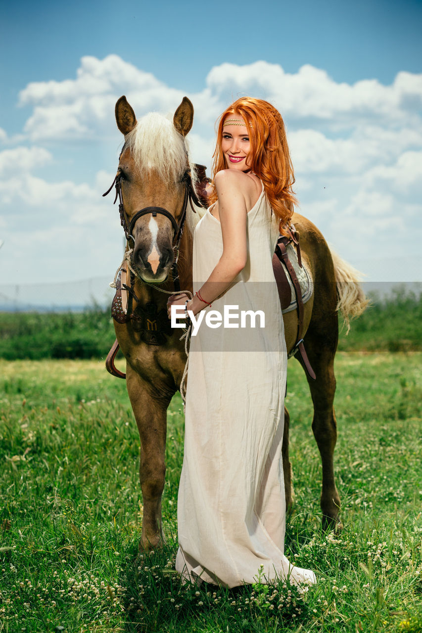 Full length portrait of beautiful woman with horse standing on grassy field against sky