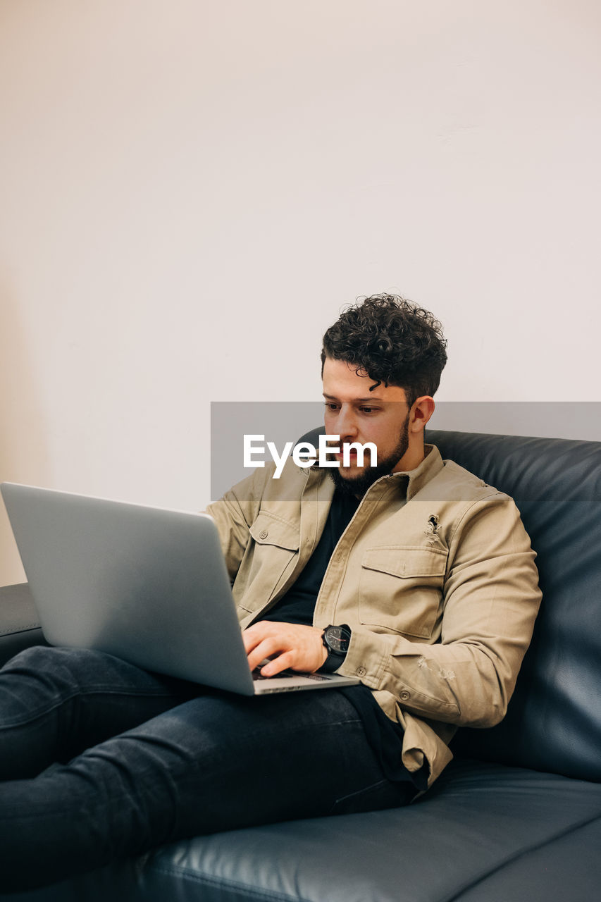 YOUNG MAN USING MOBILE PHONE WHILE SITTING ON SOFA AT HOME