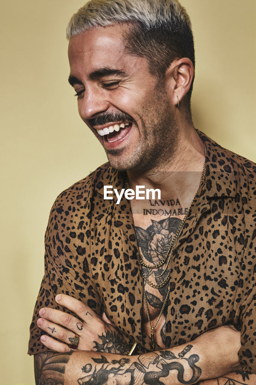 Portrait of cheerful fashionable male model with tattoos wearing trendy leopard shirt standing against beige background with closed eyes