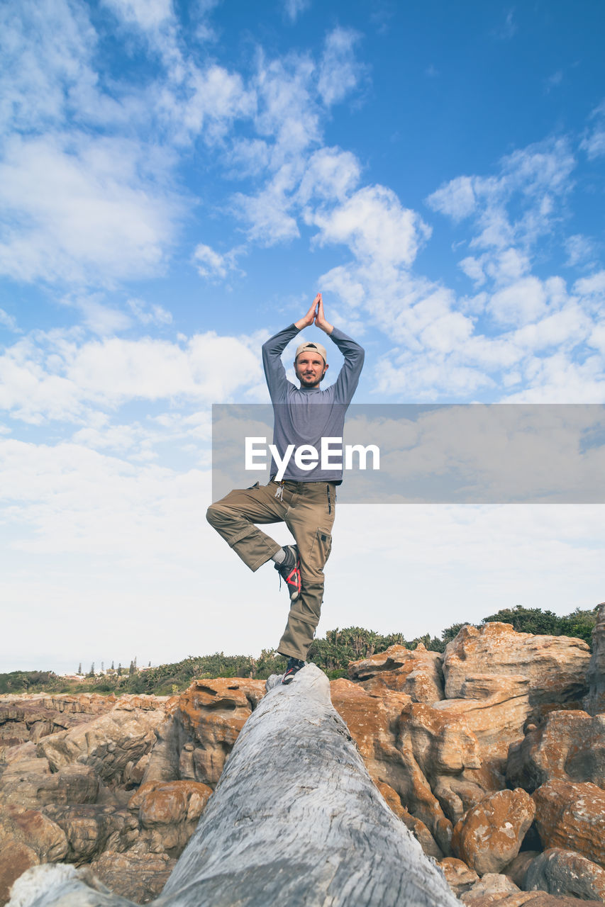 Full length of young man doing yoga on log against cloudy sky