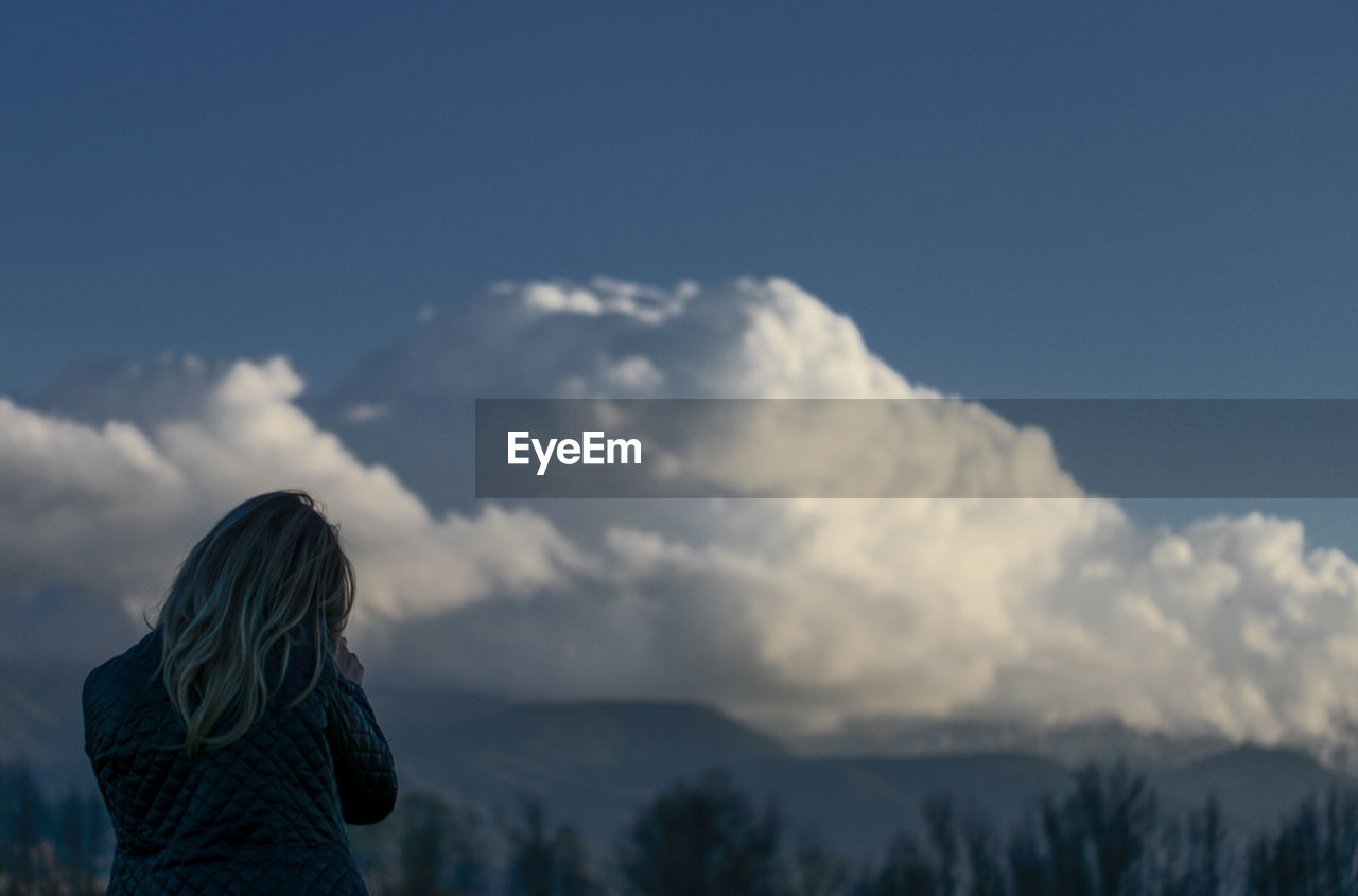 REAR VIEW OF WOMAN STANDING AGAINST CLOUDS