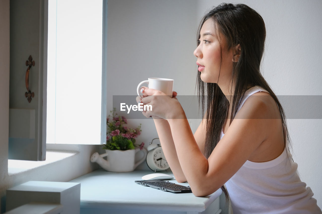SIDE VIEW OF YOUNG WOMAN HOLDING COFFEE CUP AT HOME