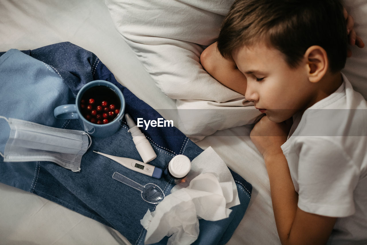 Sick child sleepping on bed. nearby there are tray with medicines, fruit drink, medical mask