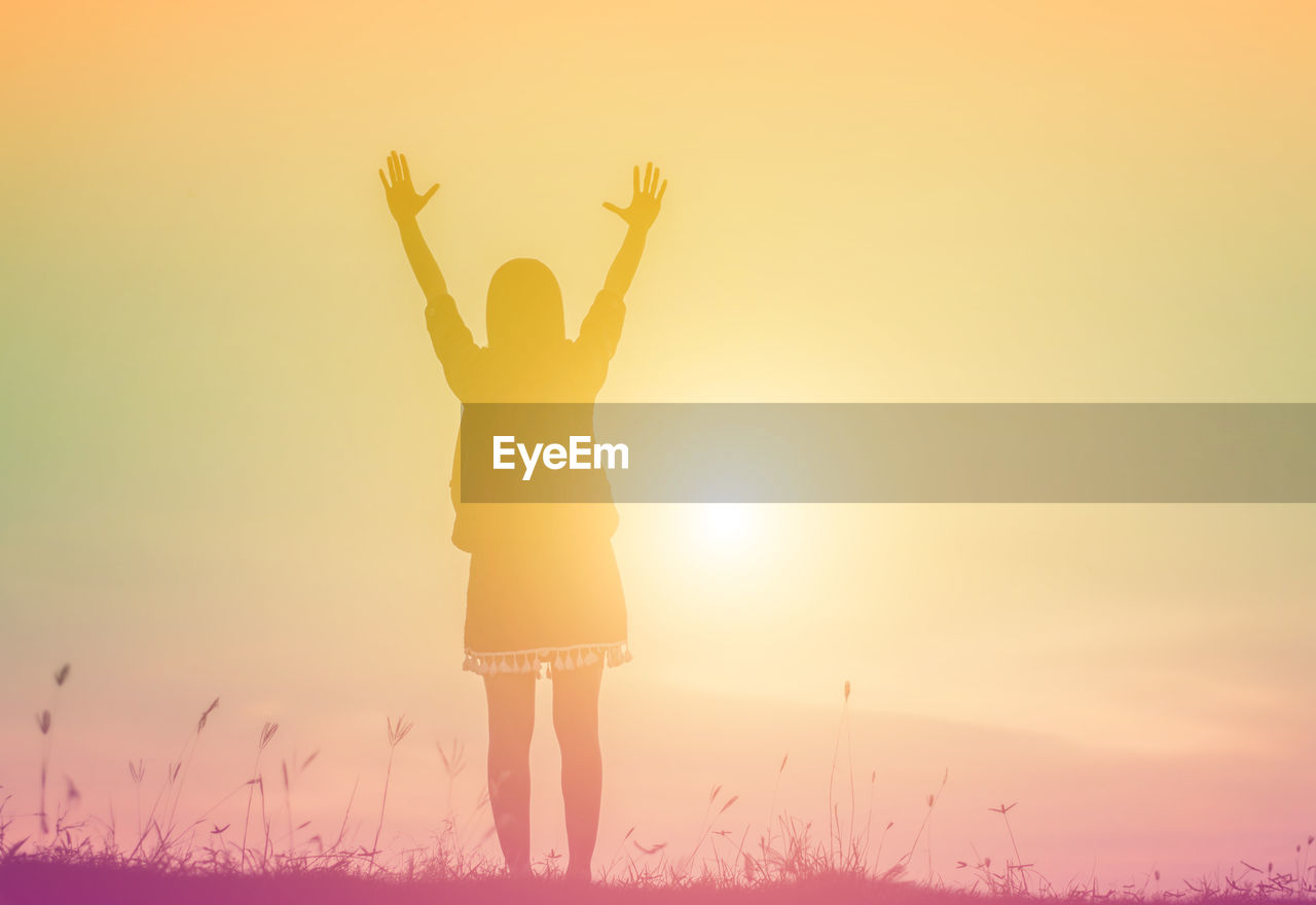 REAR VIEW OF WOMAN STANDING ON FIELD AT SUNSET