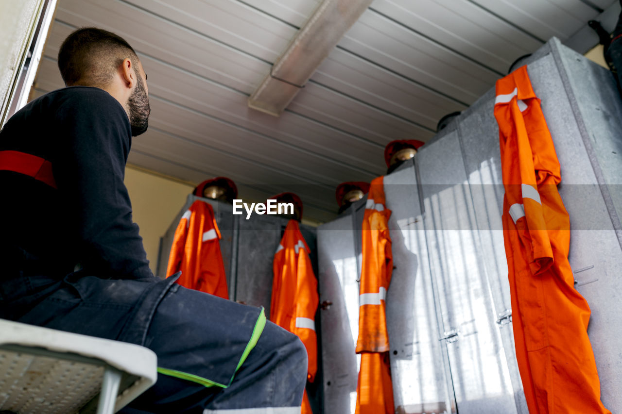 From below unknown firefighter sitting near a row of metal lockers with helmets on top and orange uniform hanging down