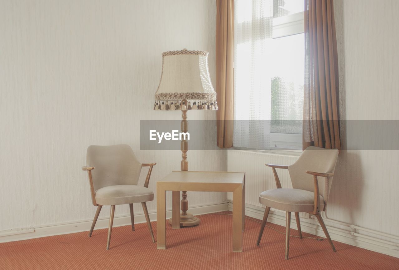 Chairs with table and electric lamp arranged by window at home