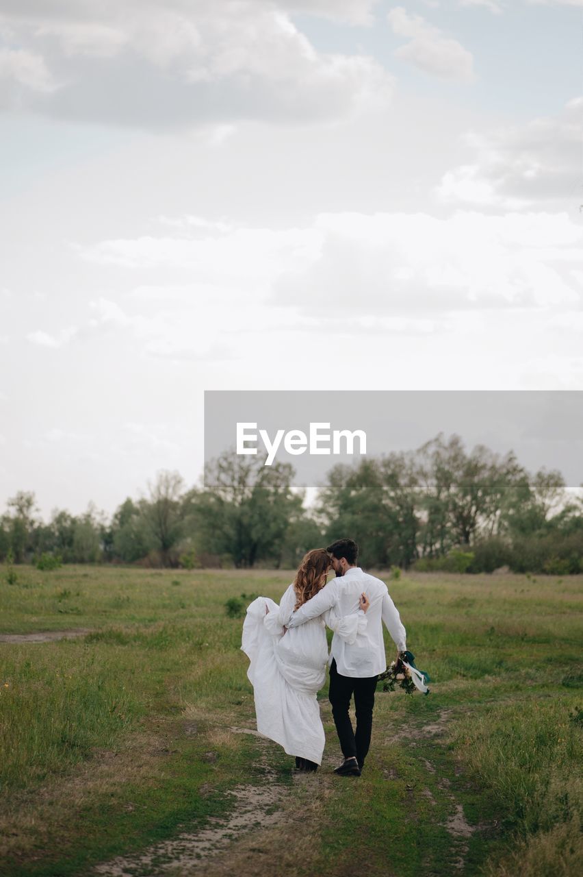 Newlywed couple walking on field against sky