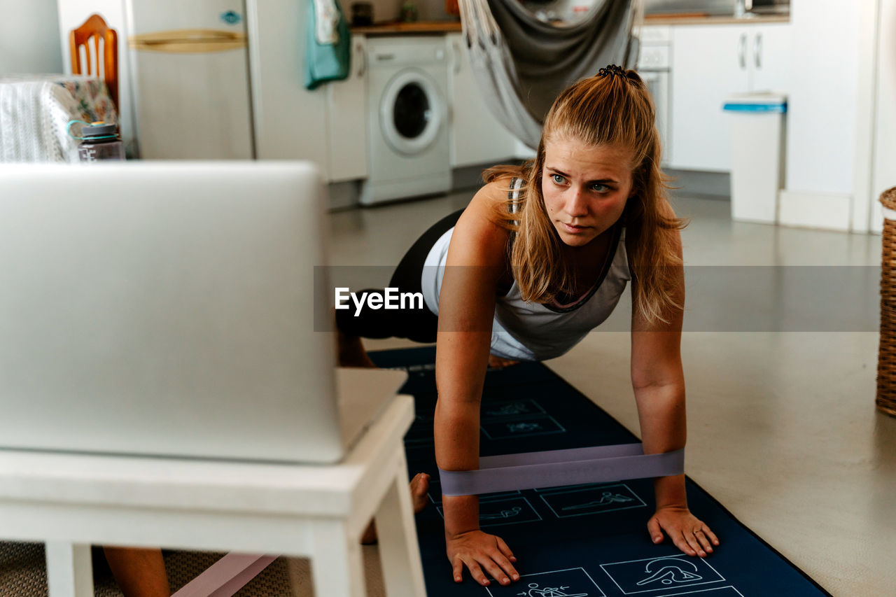 Sportswoman performing exercise with resistance bands while watching tutorials on laptop and working out at home