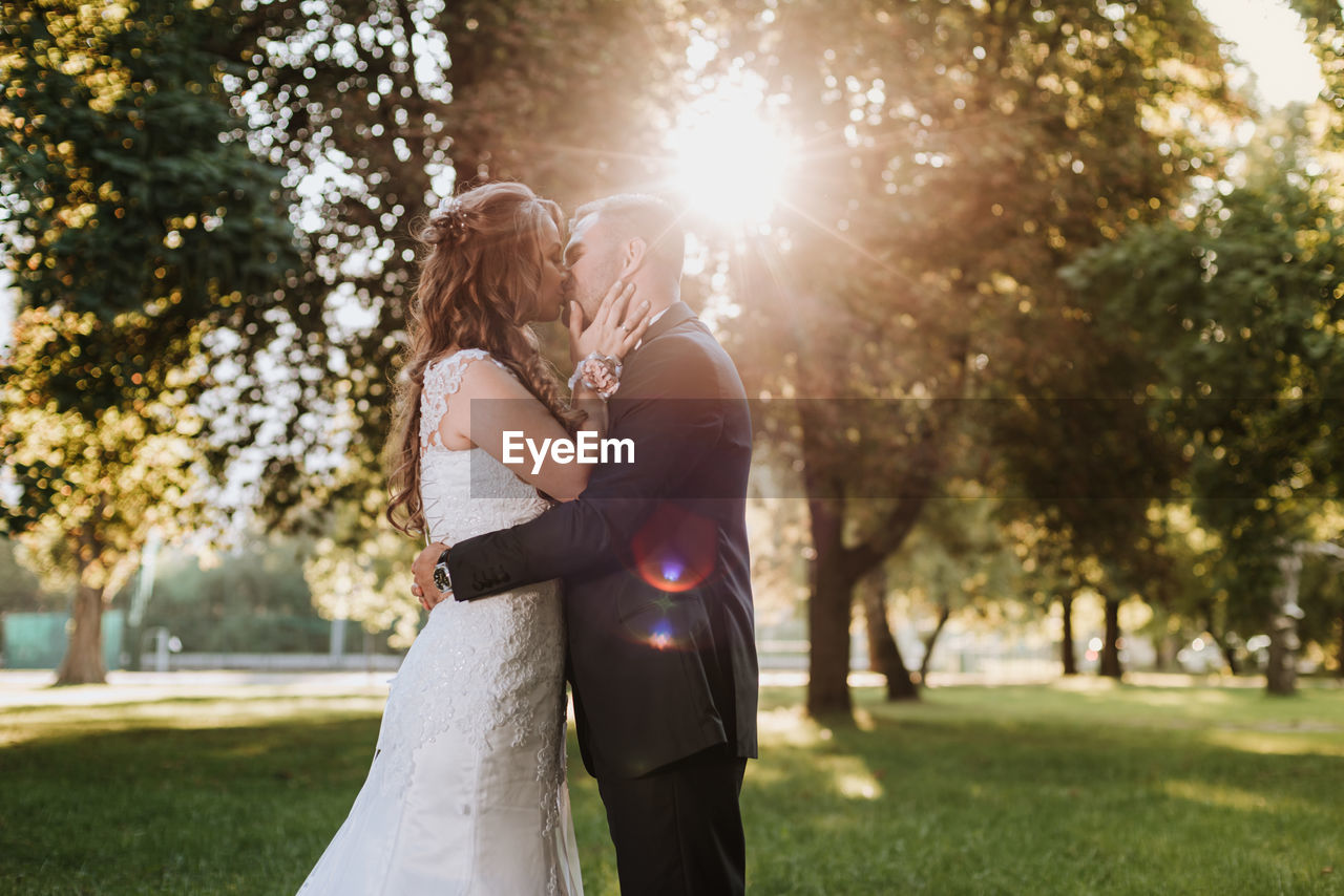 Side view of couple kissing while standing against trees