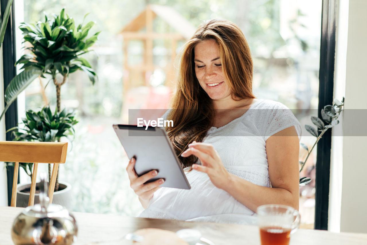 Cute freckled redhead woman is typing in a tablet computer and smiling.