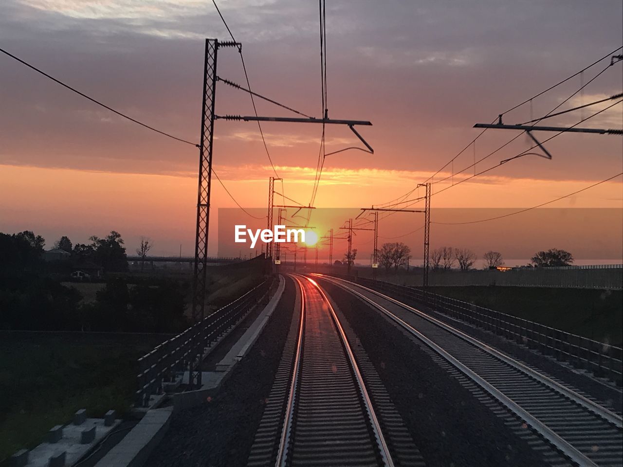 sunset, transportation, railroad track, sky, track, rail transportation, electricity, cable, mode of transportation, technology, nature, architecture, cloud, line, electricity pylon, power line, power supply, no people, travel, vanishing point, city, evening, sun, built structure, public transportation, environment, outdoors, railway, train, dusk, transport, power generation, landscape, railroad station, twilight, vehicle, dramatic sky, electrical supply, railroad station platform, orange color, sunlight, diminishing perspective, travel destinations, silhouette, business finance and industry, beauty in nature, the way forward