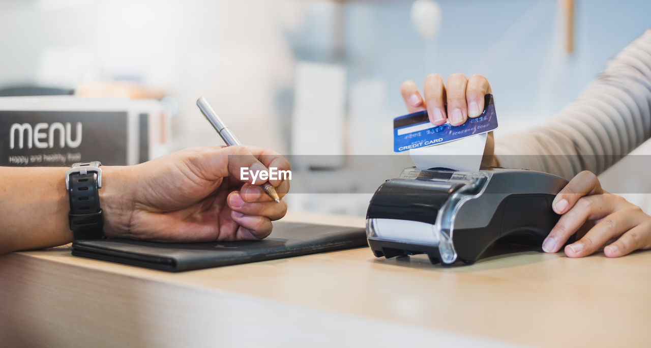 Cropped hand of person with pen while woman using credit card reader at table