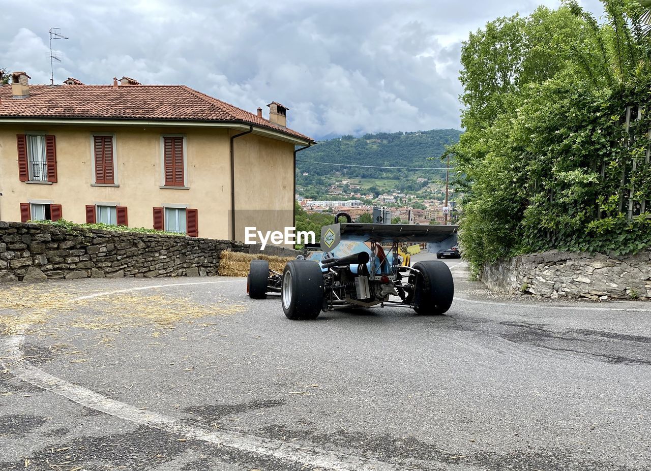 transportation, asphalt, mode of transportation, architecture, built structure, road, building exterior, cloud, car, building, wheel, nature, sky, vehicle, tire, day, land vehicle, house, city, plant, tree, street, outdoors, residential district, motor vehicle