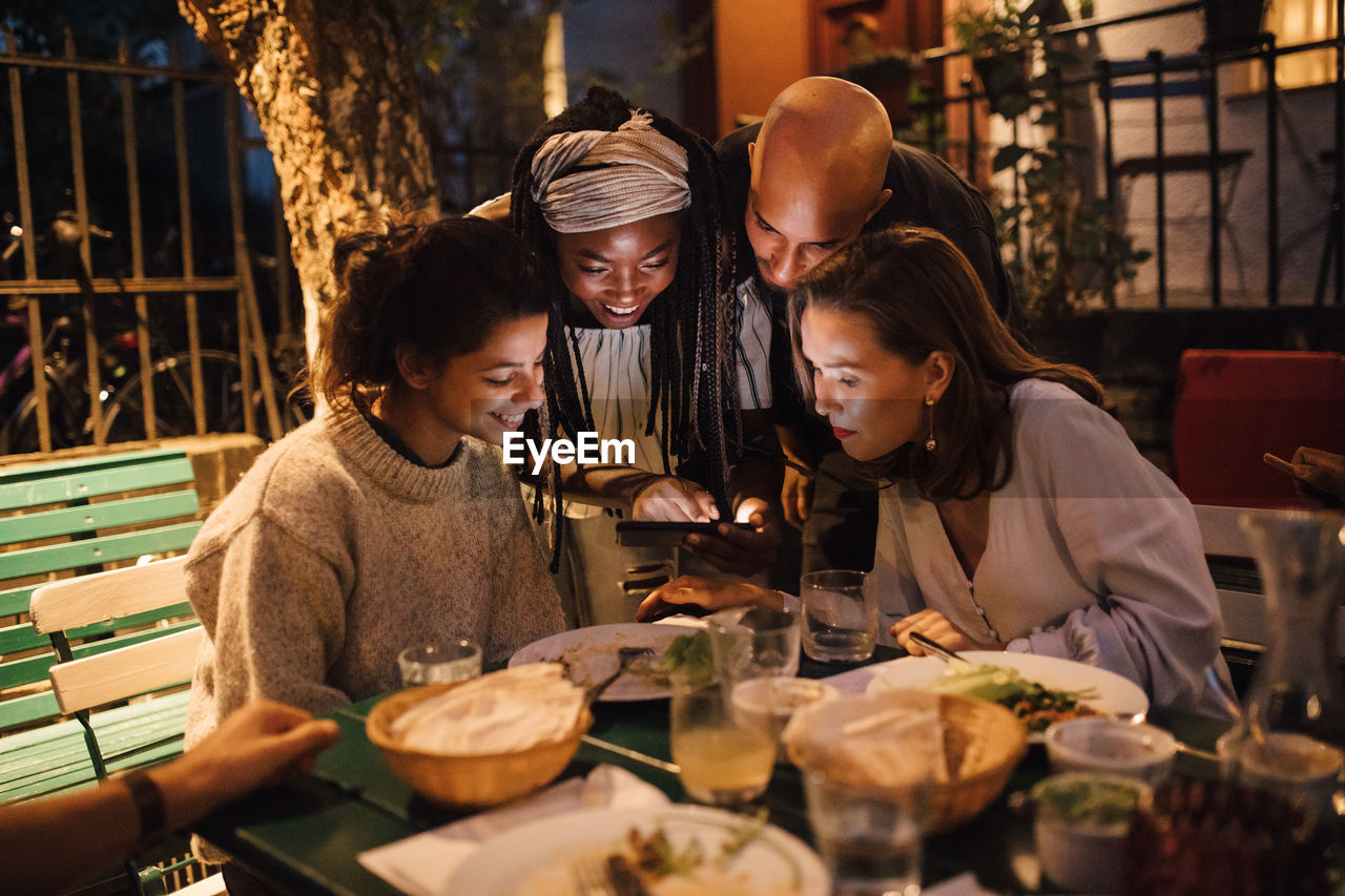 Happy young woman showing smart phone to friends at table during dinner party