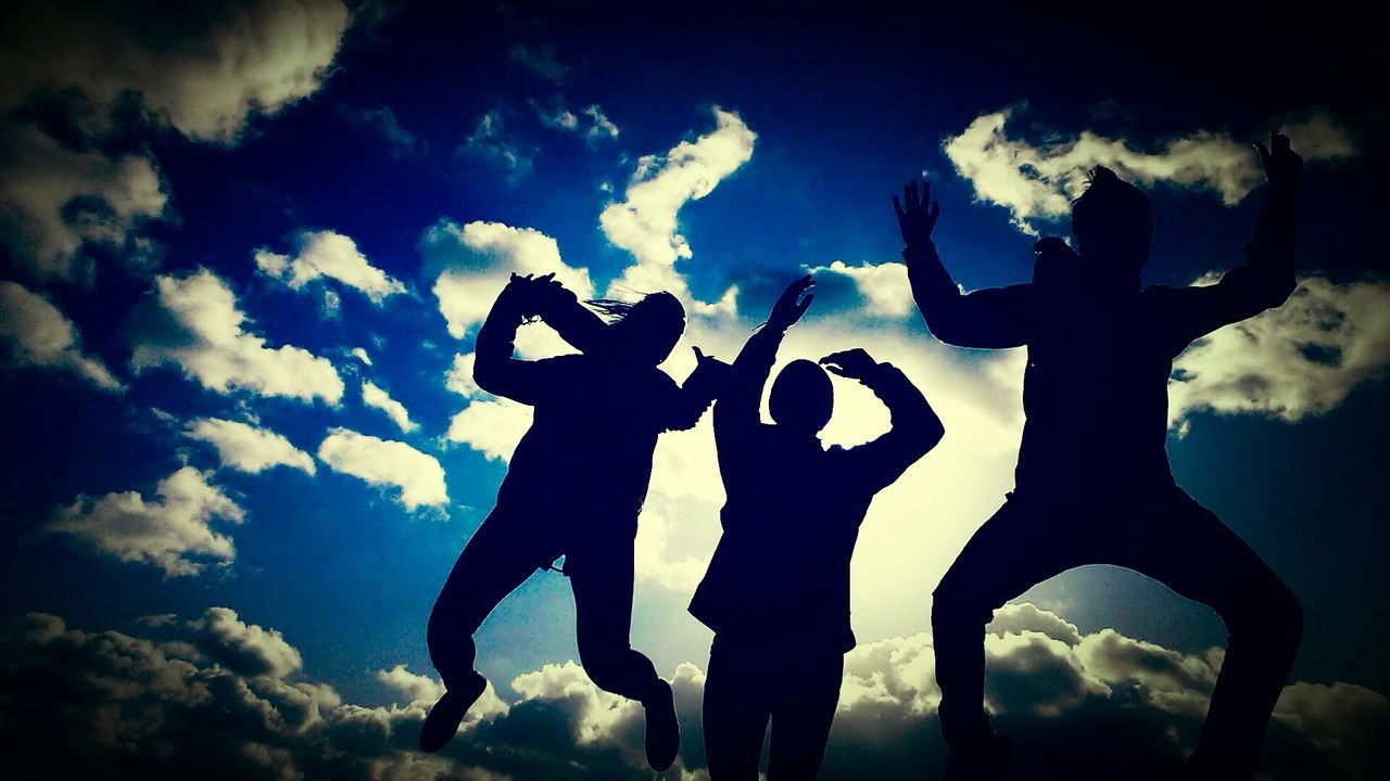 Low angle view of silhouette friend jumping against blue cloudy sky