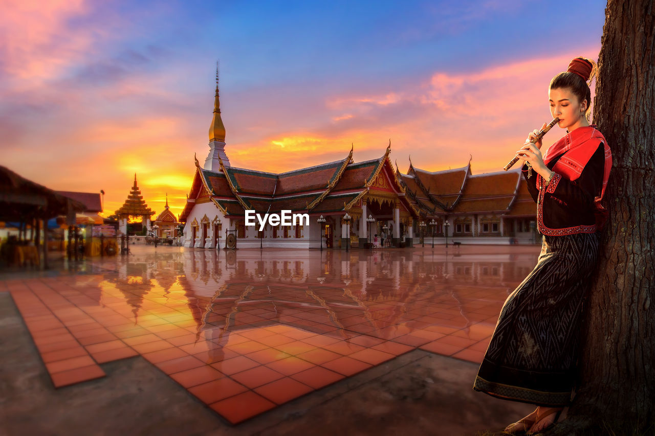 Young woman playing flute while standing against temple during sunset