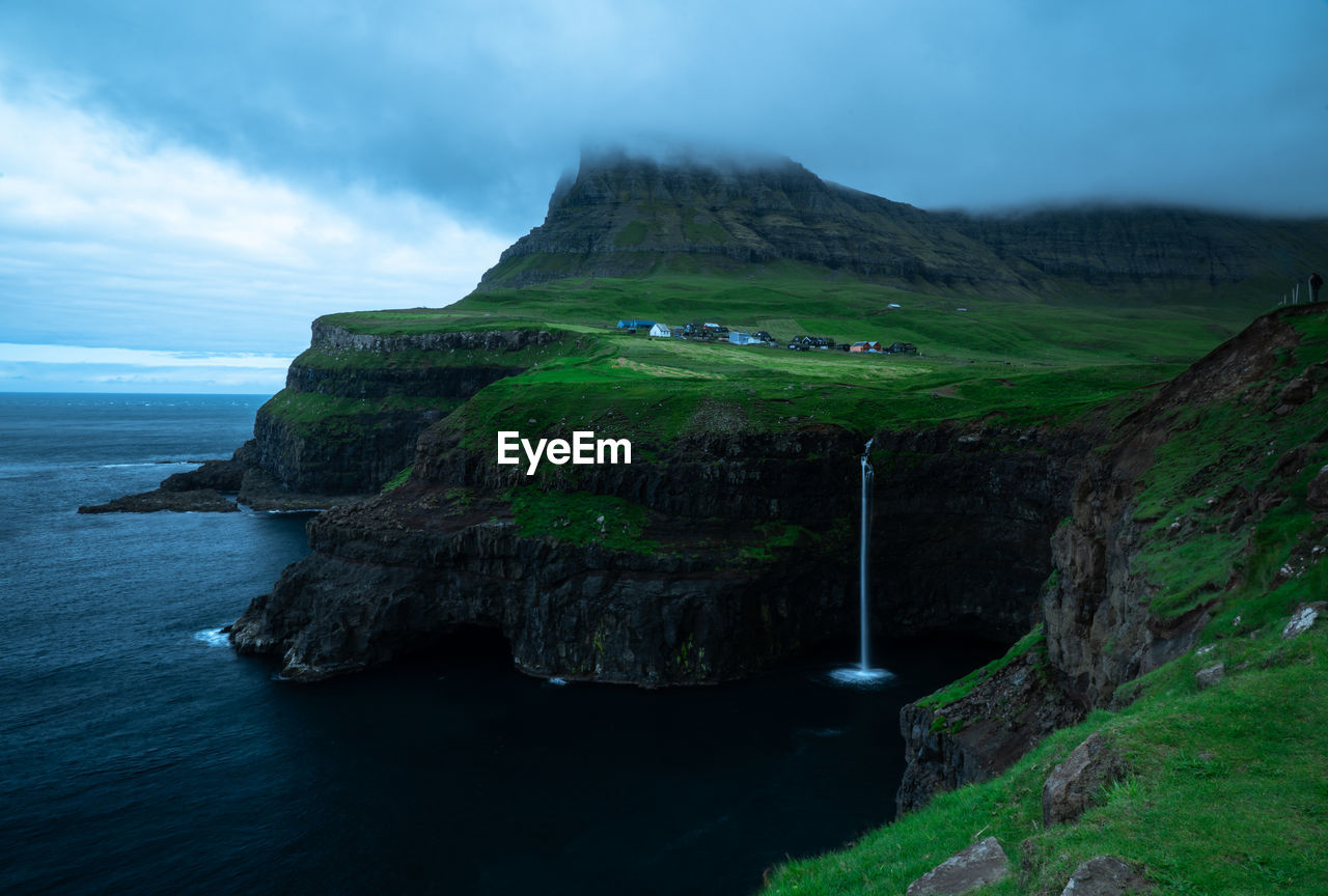 Scenic view of waterfall, green mountains and sea under a cloudy sky.