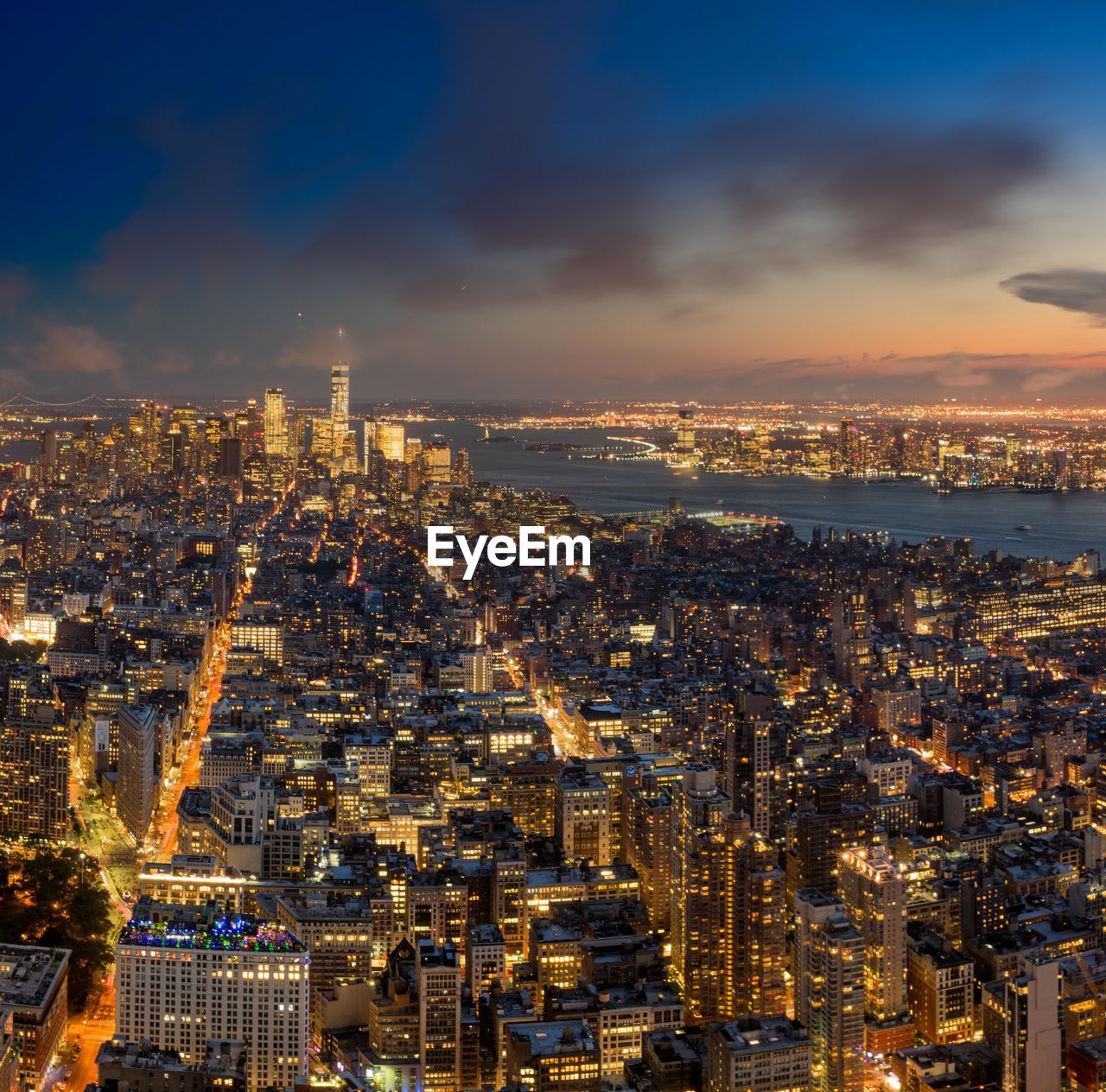 High angle view of illuminated cityscape by river against cloudy sky at dusk