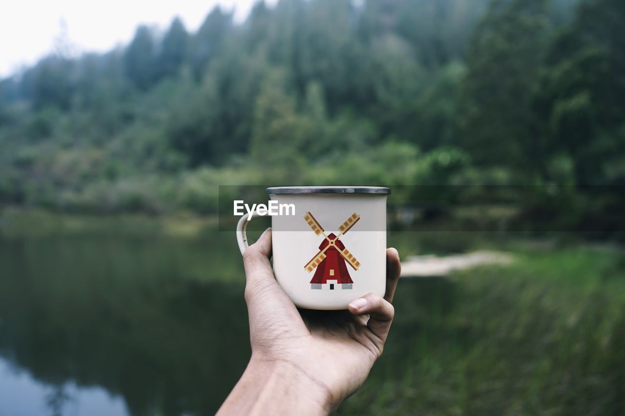 Close-up of hand holding mug over lake