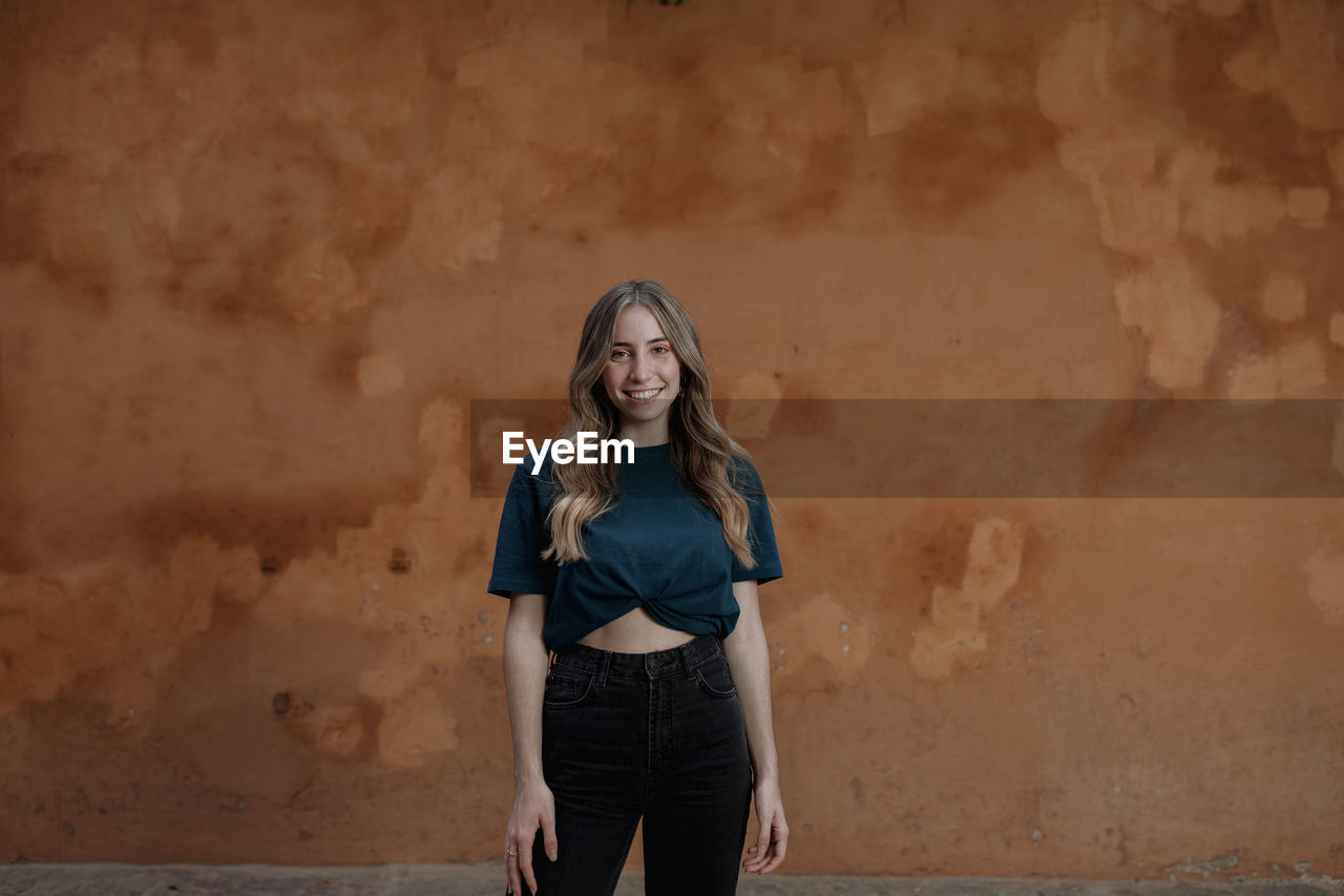 Young content blonde female in casual wear with wavy hair looking at camera while standing on brown background in town